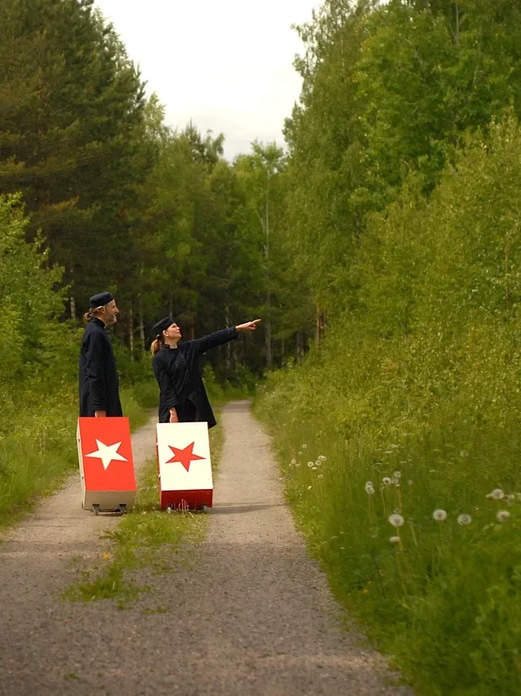Två personer går på en grusväg i en skog, båda bär svarta kläder och har långa hår, en av dem pekar mot trädtopparna. Den ena personen bär en låda med en vit stjärna på, den andra med en röd stjärna på.