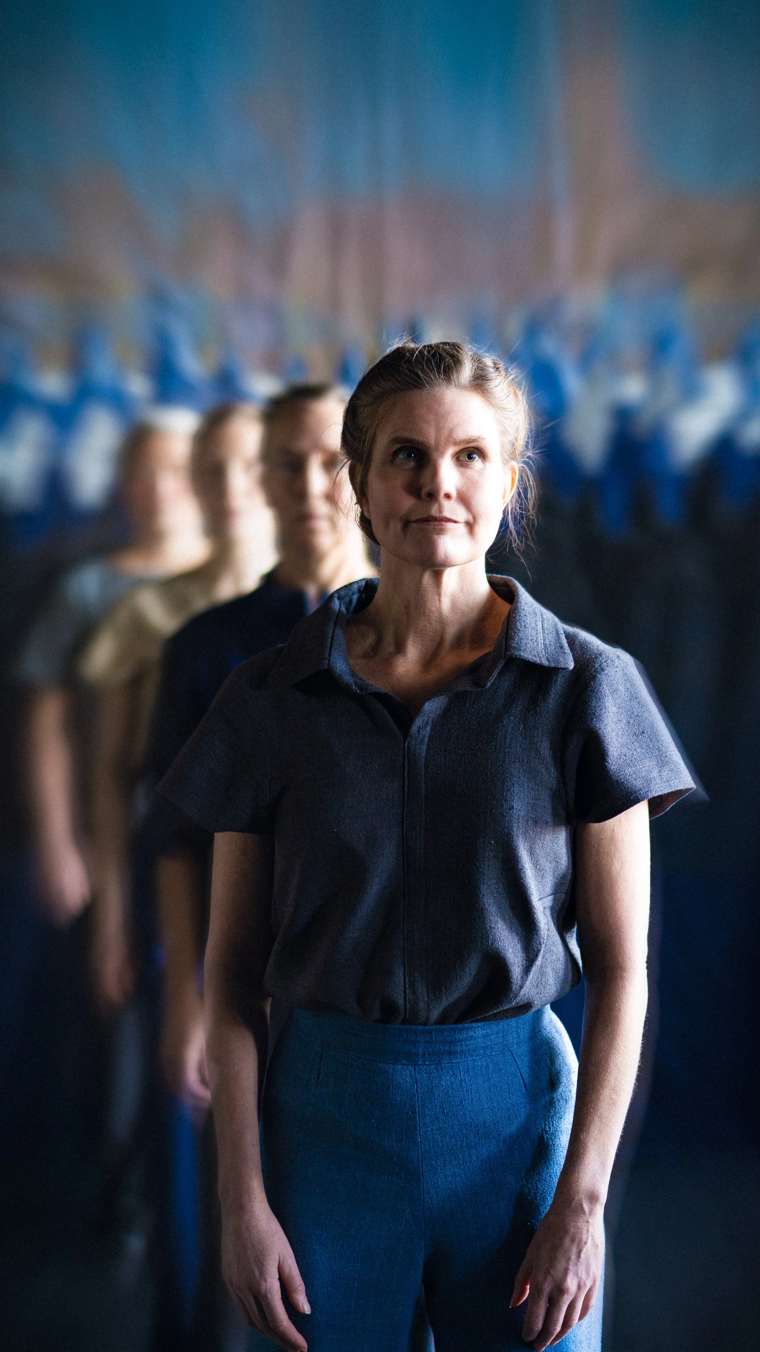 Kvinna står framför en grupp personer, alla i samma posering, i en konstnärlig bild med fokus på kvinnan i förgrunden.