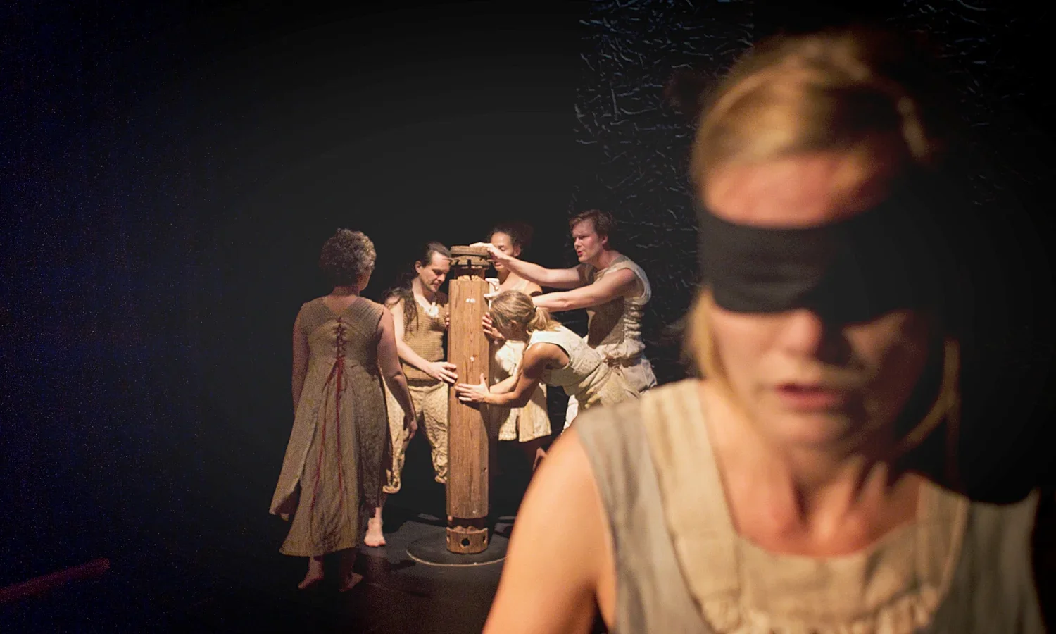 Scen från en teaterföreställning med fem barfota personer i bakgrunden som arbetar tillsammans på ett trästycke, medan en kvinna i förgrunden är blindfoldad.