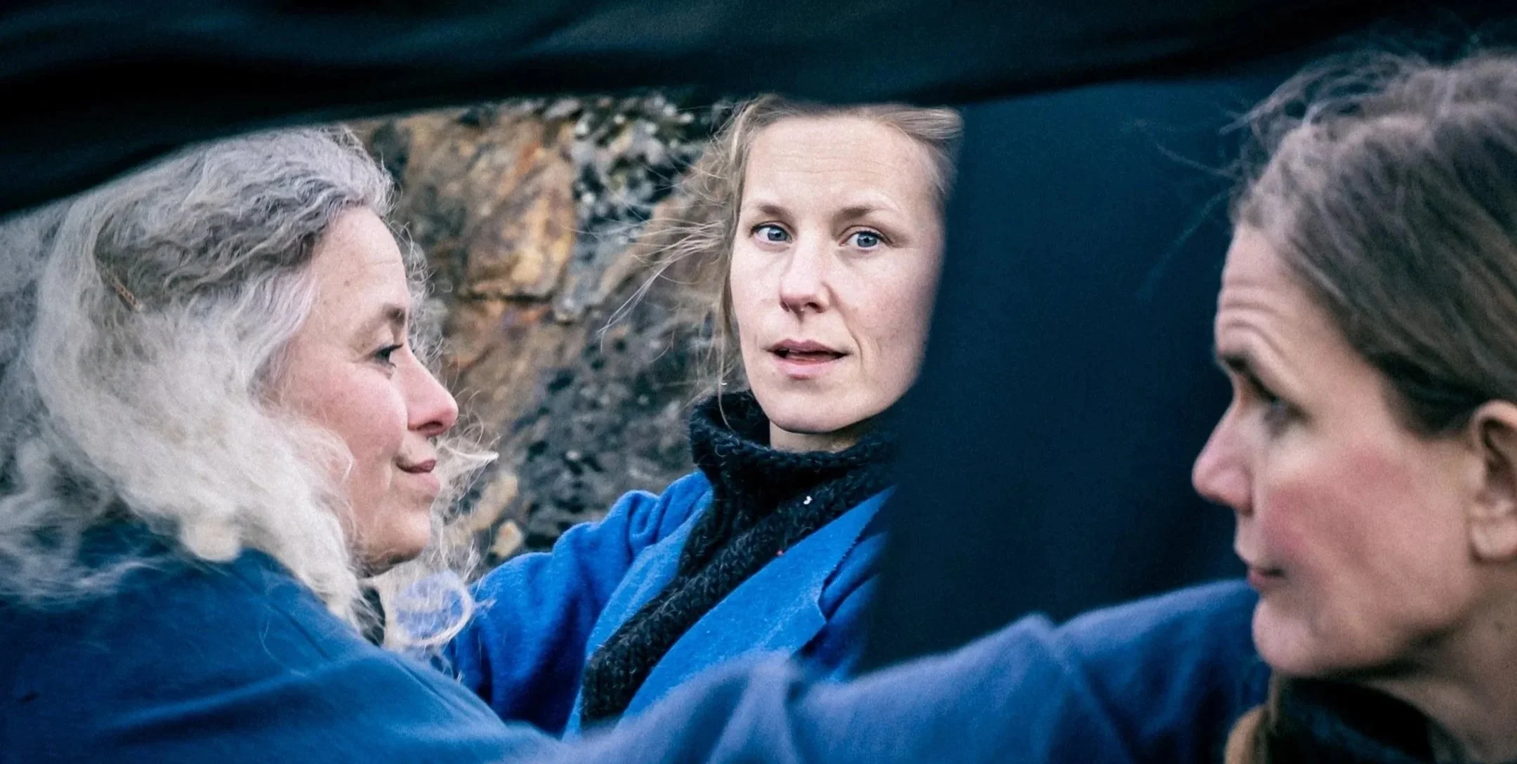 Tre kvinnor i samtal utomhus, omgivna av natur och stenar.