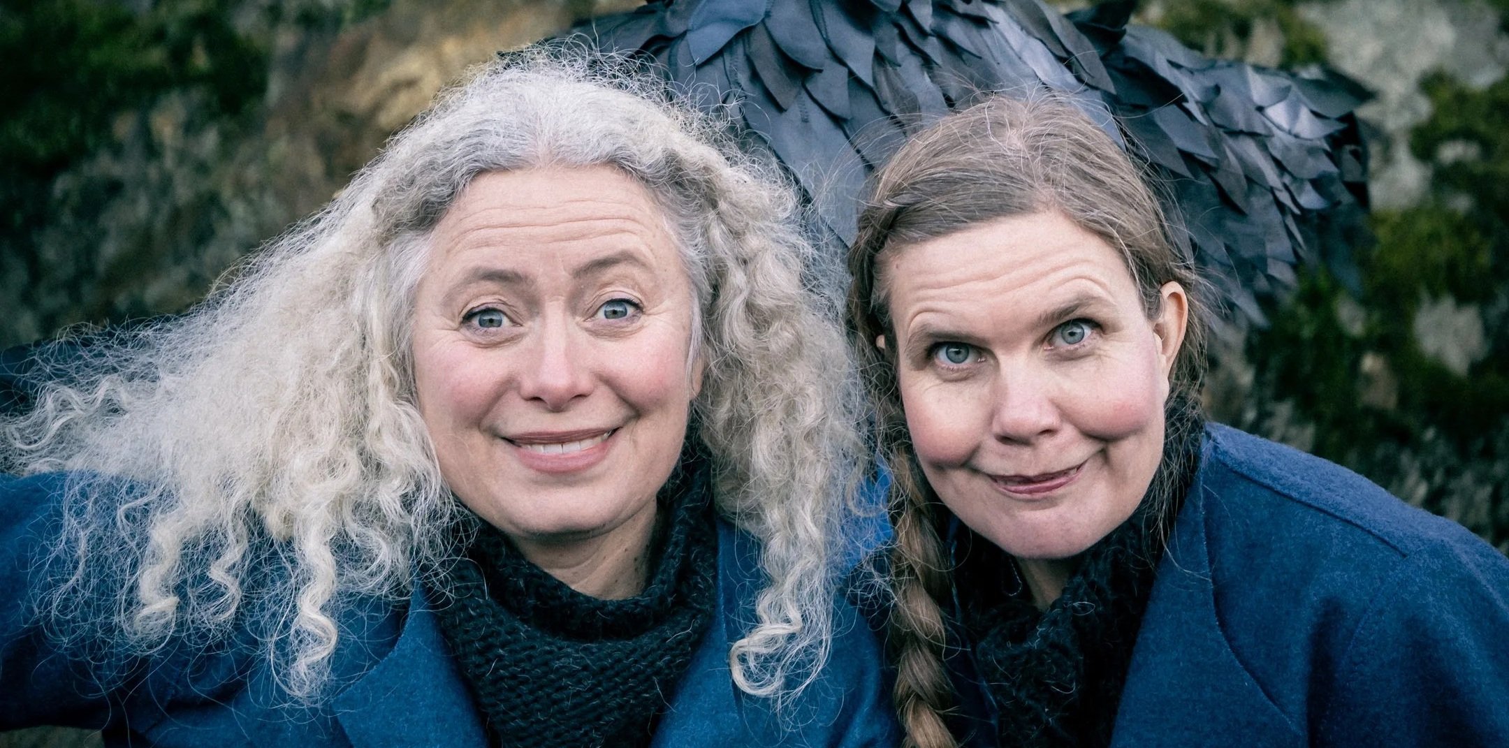 Två kvinnor med blå kläder och mörka tröjor, står nära varandra utomhus, båda ler och tittar mot kameran.