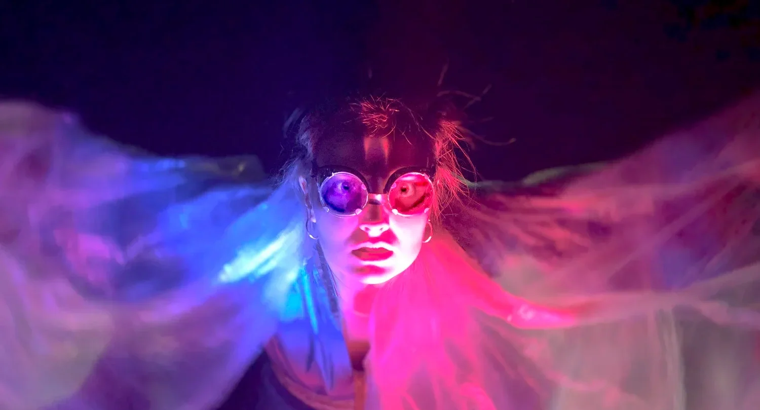 En person med "goggles" som tittar rakt in i kameran, ståendes med armarna utsträckta i rosa och blå ljus, i ett mörkt rum.
