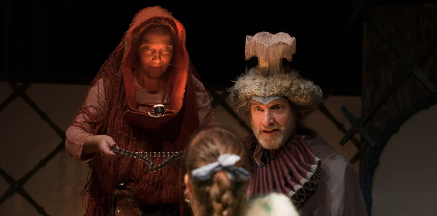 Två personer, en äldre kvinna och en man med 1600-tals kläder. De är inomhus i en teatermiljö.