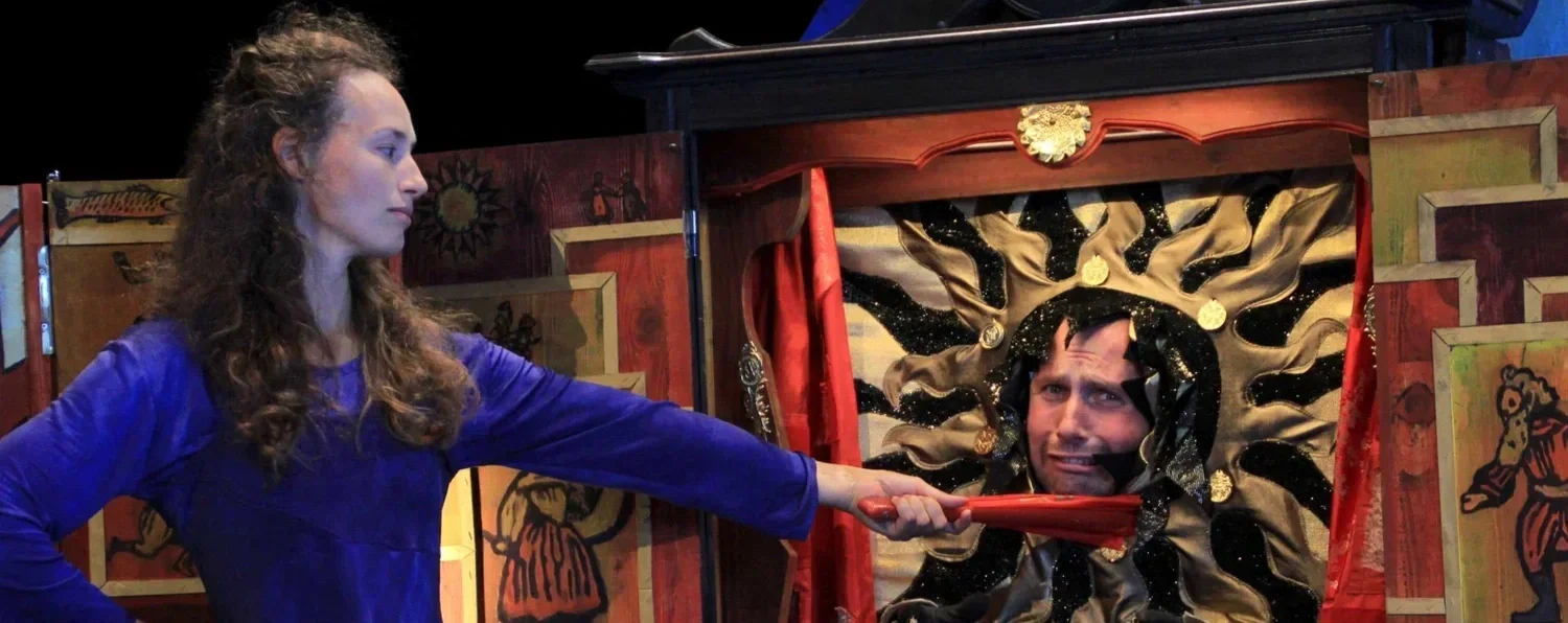 En kvinna i blå klänning som håller en pinne mot ett fäste i ett teaterdockspel.