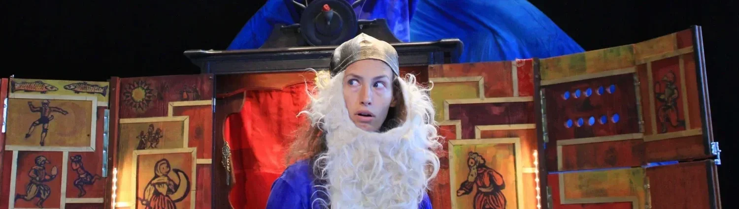 En person klädd som en jultomte med vitt skägg och en tomtedräkt, framför en scen med färgglada dekorationer.