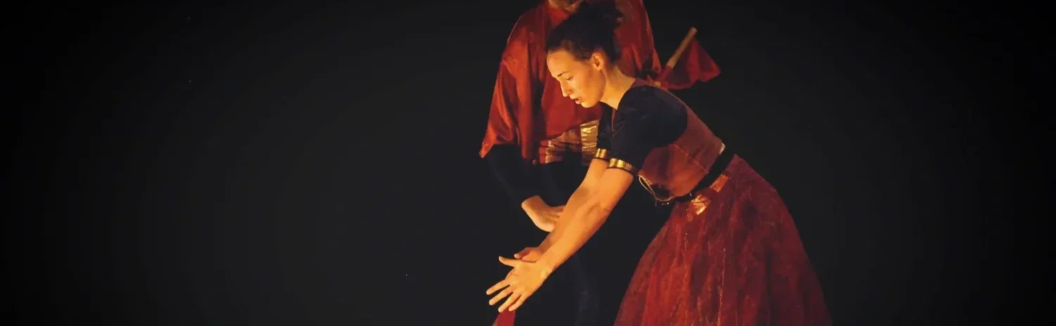 Två personer i teaterkostymer under en föreställning. En kvinna böjer sig framåt medan en annan person står bakom henne.