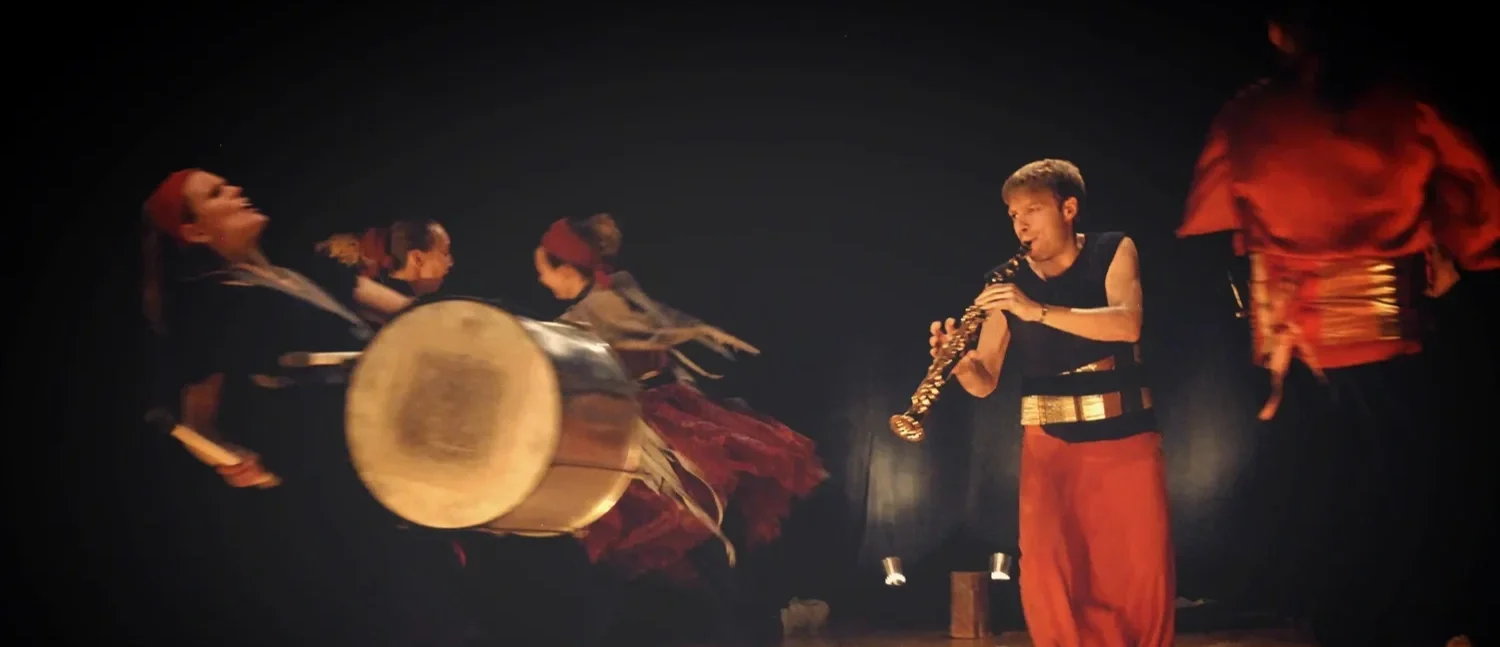 Människa som spelar klarinett omgiven av dansare i rörelse, med mörk bakgrund.