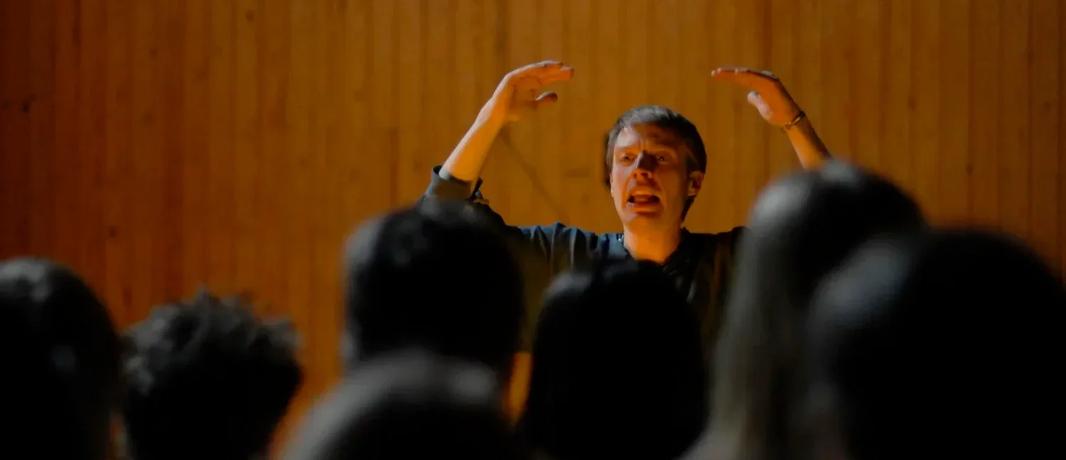 En person framför en publik, som verkar vara en lärare eller talare, gestikulerar med händerna medan hen talar, mot en trävägg.