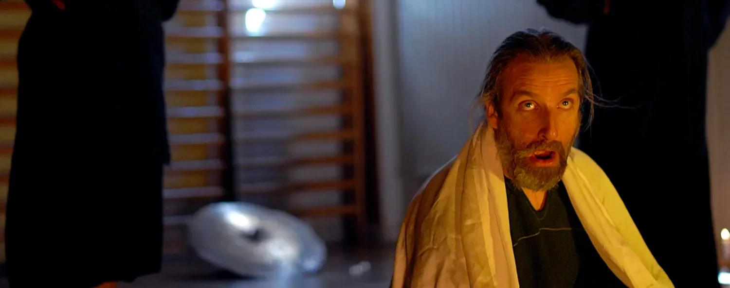 Man med långt hår och skägg som sitter på golvet, ser förvånad ut, inomhus med träväggar och en ljuskälla.