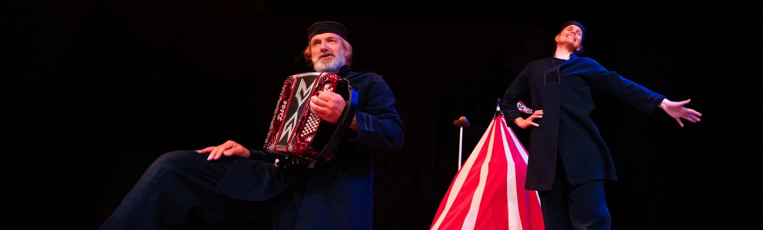 En man och en kvinna som uppträder på en scen, med en akordeon och en clownfigur i bakgrunden.