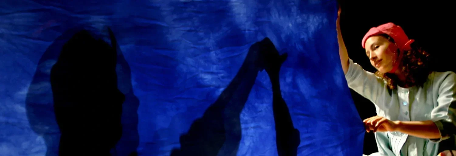 En flicka med röd keps och ljus tröja håller i en blå tygbit framför en ljuskälla, vilket skapar skuggbilder av hennes händer och kropp på tyget.