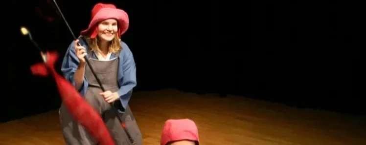 En kvinna som ler, klädd i en blå jacka och en rosa hatt, håller i en fiskespö i ett rum med trägolv och mörk bakgrund.