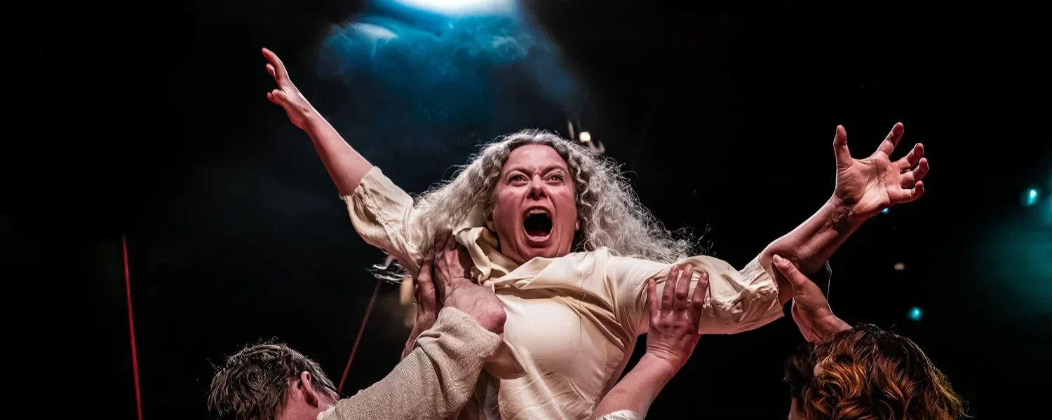 En kvinna med grått lockigt hår skriker med armarna utsträckta medan två personer håller henne uppe på en mörk scen.