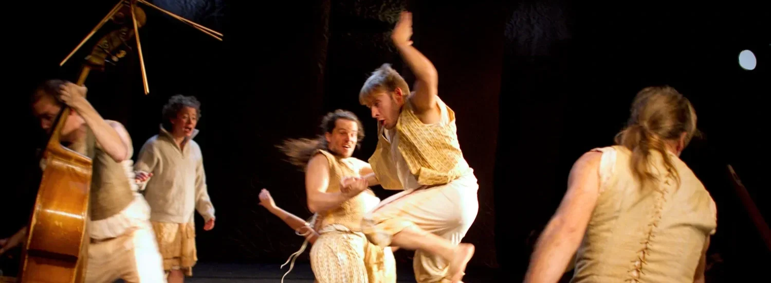 Flera personer som dansar, leker och spelar musikinstrument på en scen, klädda i beige kläder.