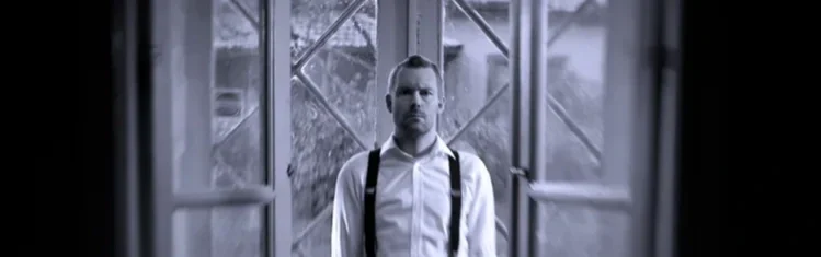 Man står i ett rum framför fönster, klädd i vit skjorta och svarta hängslen.