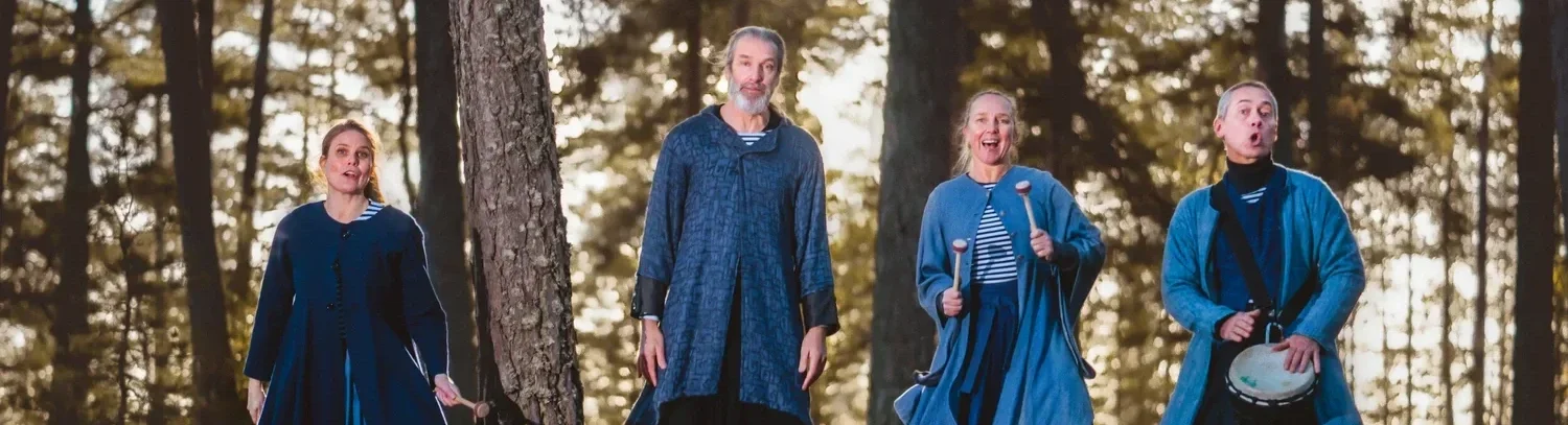 Fyra personer står i en skog, alla är klädda i blå kläder och verkar sjunga eller spela musik tillsammans.