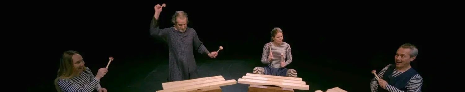 Fyra personer på scen, tre av dem håller trumpinnar, alla sitter vid lådor eller trummor, framför ett musikaliskt framträdande.