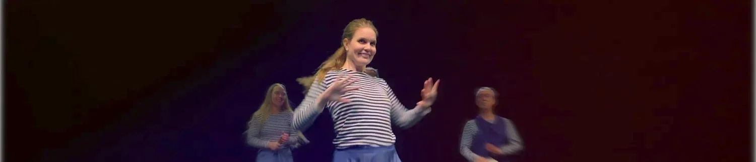 Tre kvinnor i randiga tröjor och blåa byxor som dansar på en scen mot mörk bakgrund.