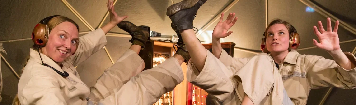 Två kvinnor i beige dräkter och hörlurar gör akrobatiska positioner inomhus.