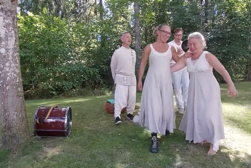 Fyra personer i vita klänningar och en person i vita kläder som går på gräset i en skogsmiljö, med ett instrument på marken till vänster.