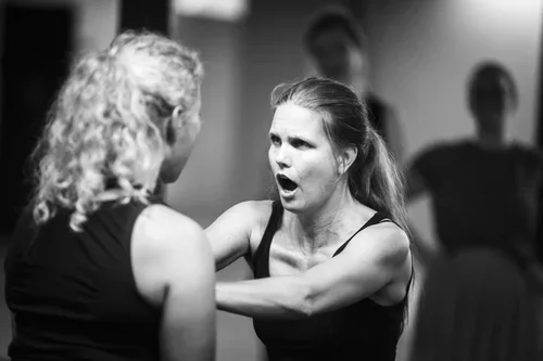 En kvinna med ett bestämt ansiktsuttryck som konfronterar en annan kvinna, med personer i bakgrunden.