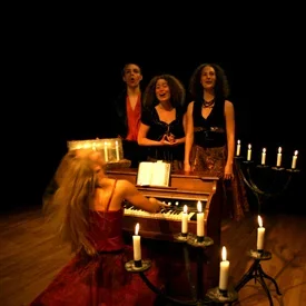 Tre kvinnor sjunger när de spelar piano, medan en annan kvinna spelar piano med tända ljus runt sig i ett mörkt stämningsfullt rum.