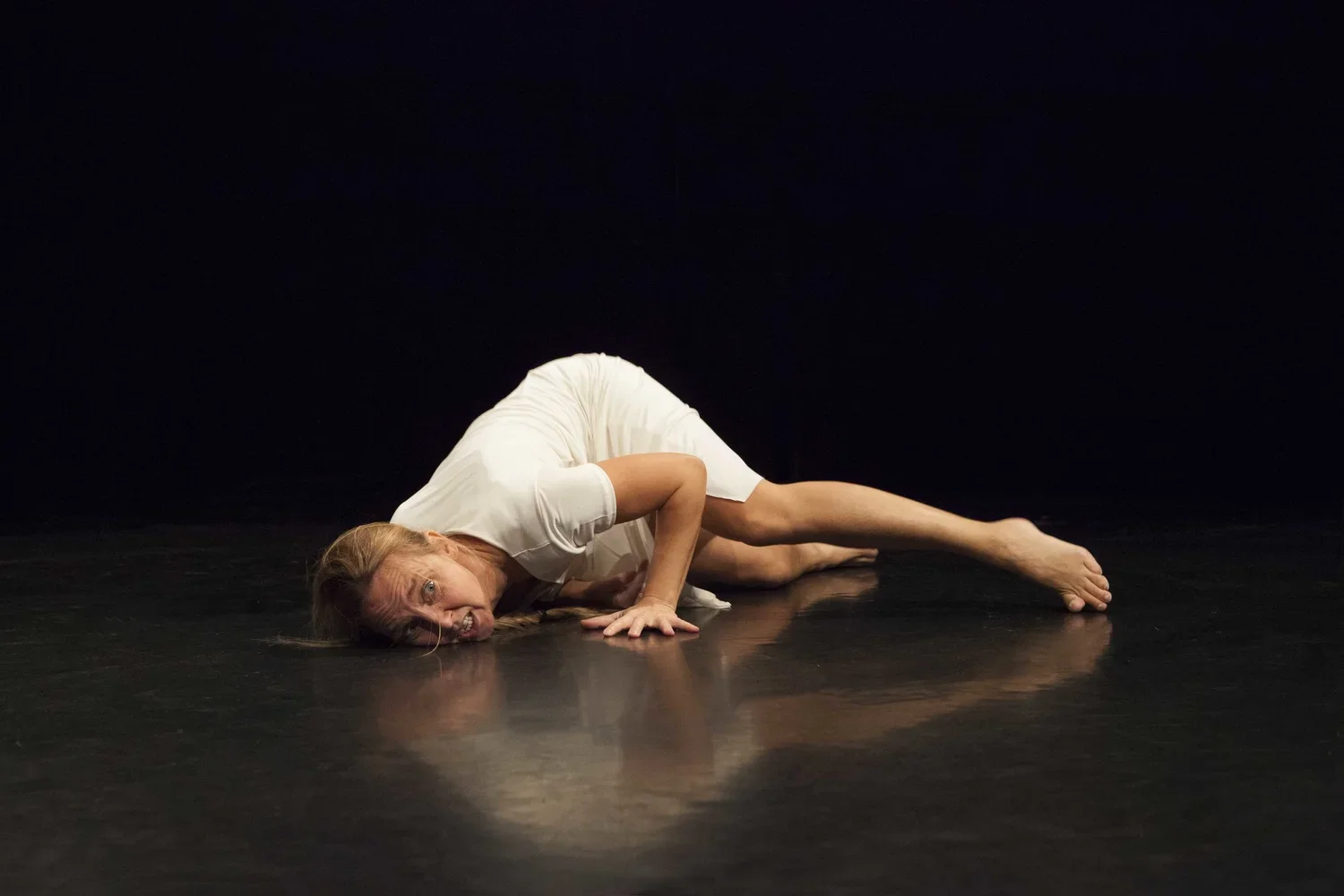 En person i en vit klänning ligger på scenen i en dramatisk pose med ansiktet nära golvet, mot en svart bakgrund.