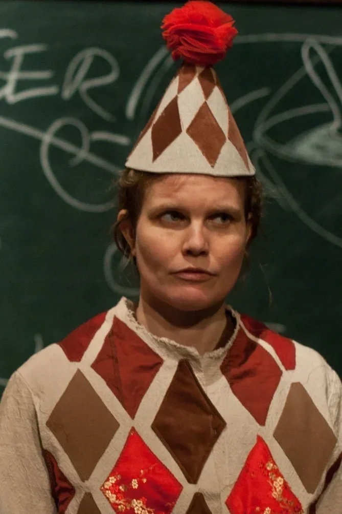 En person som bär en hatt med ett rött pompomboll och ett tröja med ett diamantmönster i beige, brunt och rött, stående framför en chalkboard med skrivna diagram.