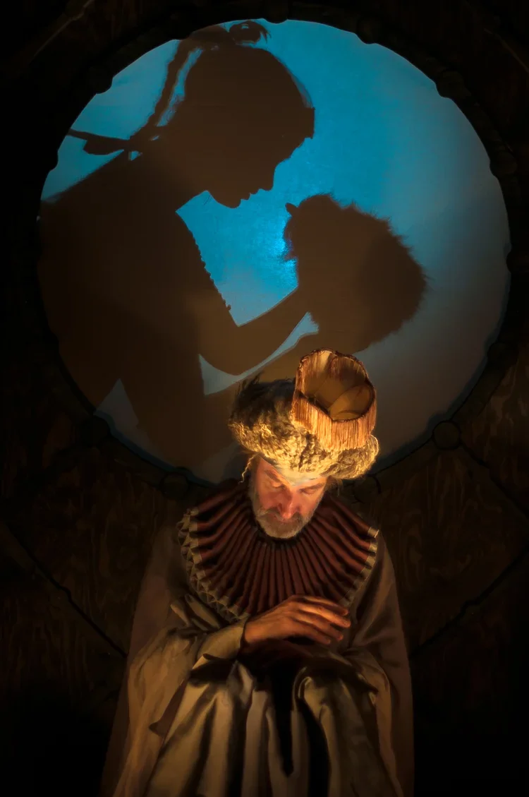 En man iklädd periodkostym med stora krås och huvudbonad, stående framför en vägg med en stor rund projicerad silhuett av en person.