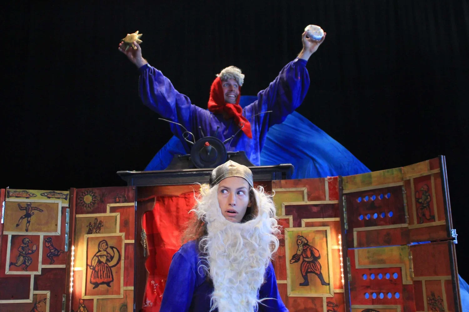 Två personer i kostymer som föreställer en kung och en narr i en teatralisk scen. Kungen står bakom en modell som är pyntad med tecknade figurer, medan narren står framför, med ett förvånat uttryck.