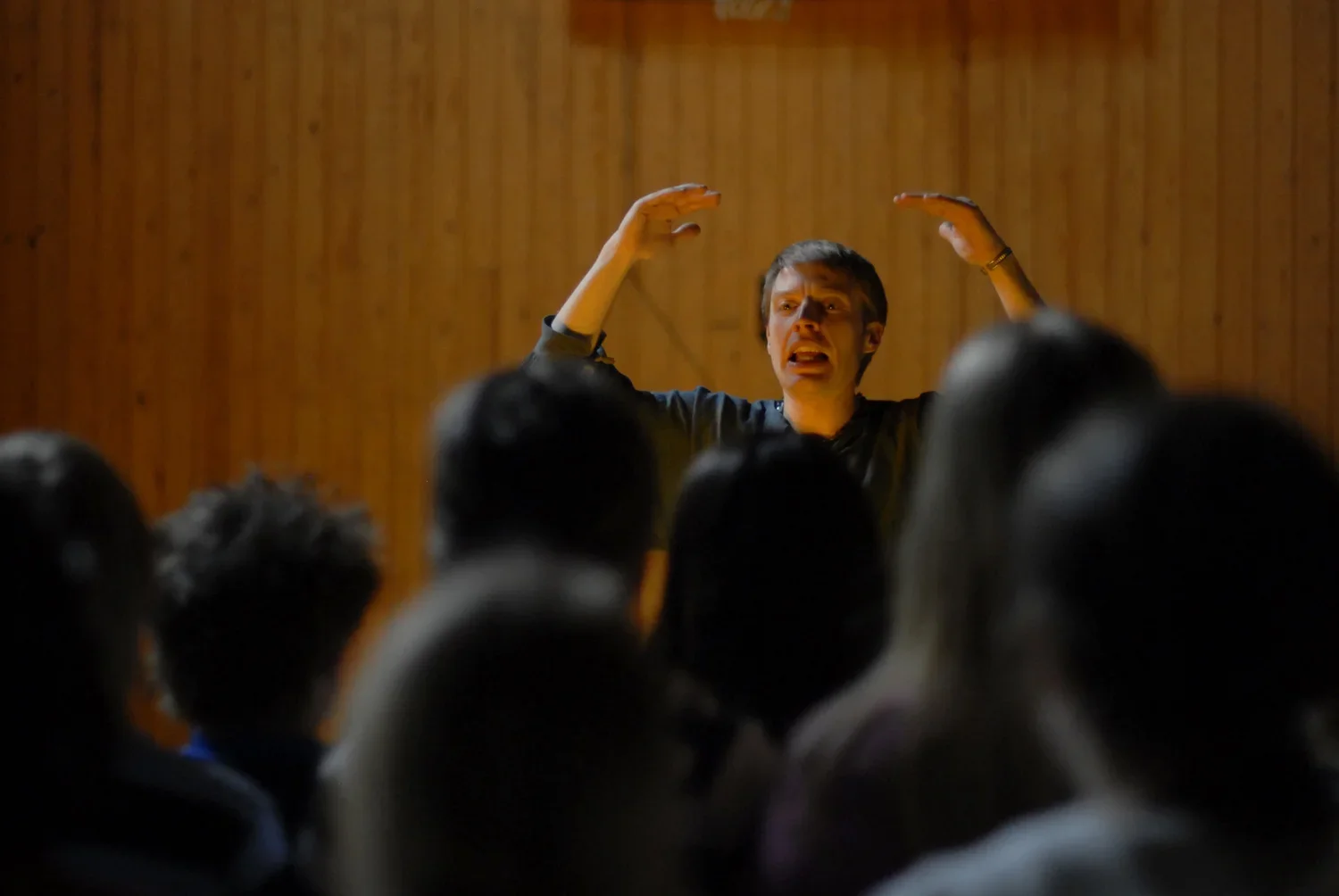 En man som talar till en publik i ett rum med träväggar, med händerna höjda i luften.