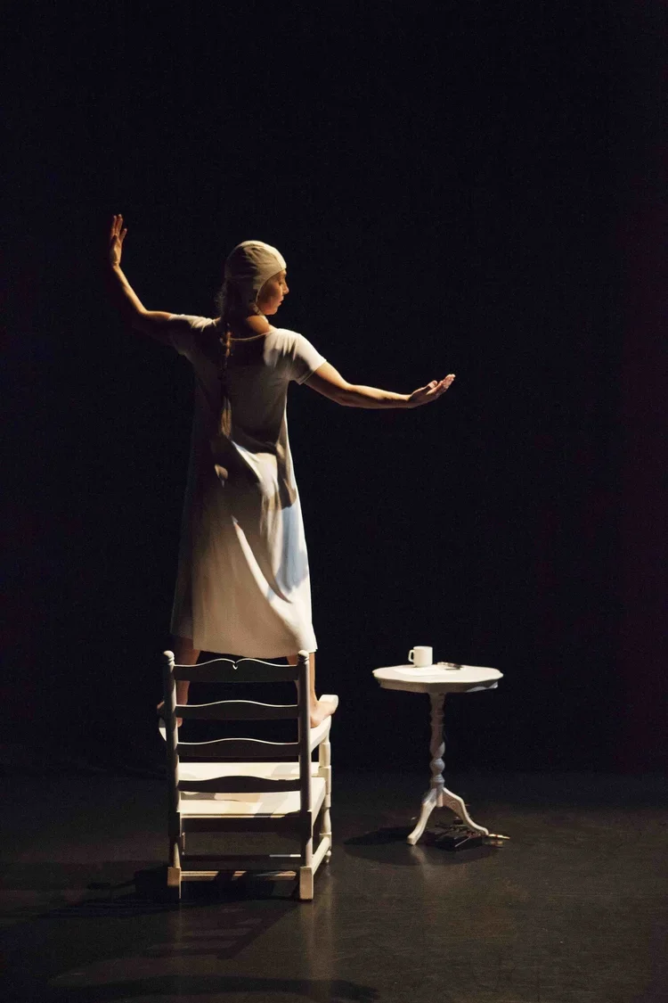 En kvinna står på stolar och bord i ett mörkt rum, med armarna utsträckta och huvudet vänt åt sidan, som om hon dansar eller utövar drama.
