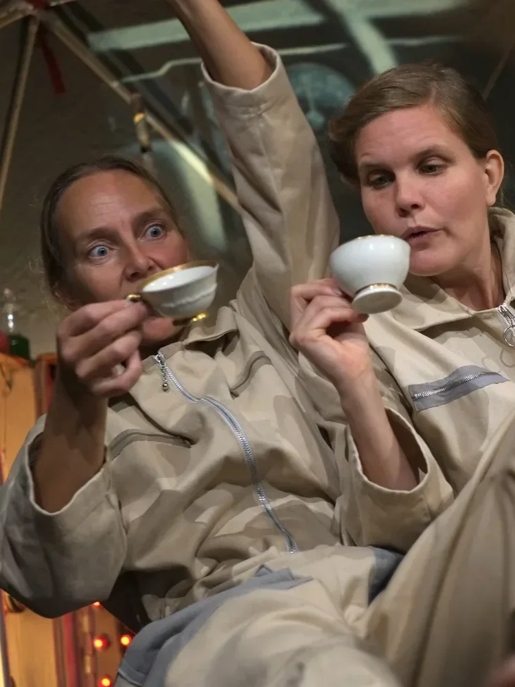 Två kvinnor i beige jumpsuits som dricker te eller kaffe ur små vita koppar.