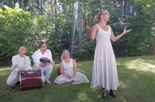 En kvinna i vit klänning som står och pratar framför en grupp av tre personer som sitter på gräset i en skogsmiljö.