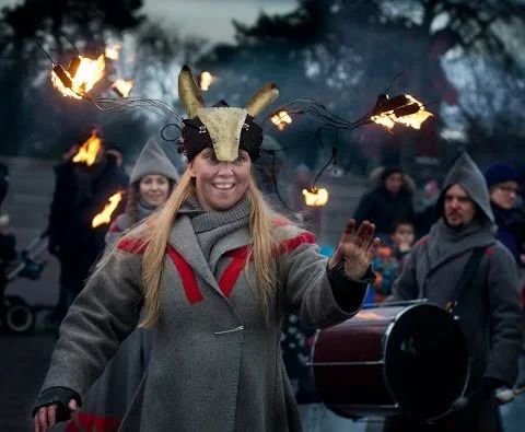 Kvinna i kostym med horn och ljusdetaljer i huvudet, omgiven av folk bärandes på eldfacklor, i ett utomhusarrangemang på kvällstid.