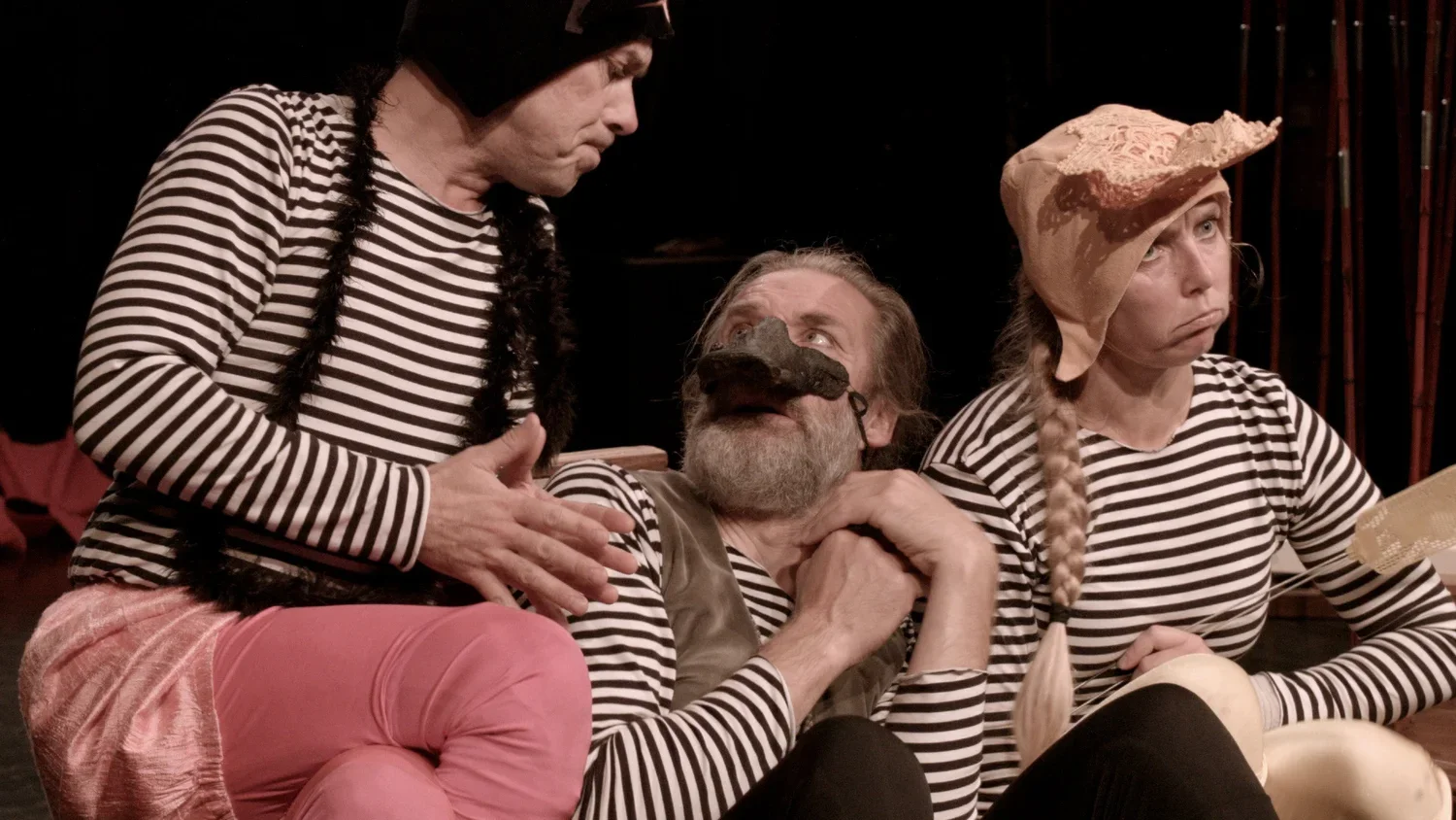Tre personer i randiga tröjor deltar i en teaterscen. En person med skägg bär en mask som täcker näsan. En kvinna med lång fläta och rosa hatt sitter intill. En person med svart mössa har ett samtal med personen som sitter mitt i bilden.
