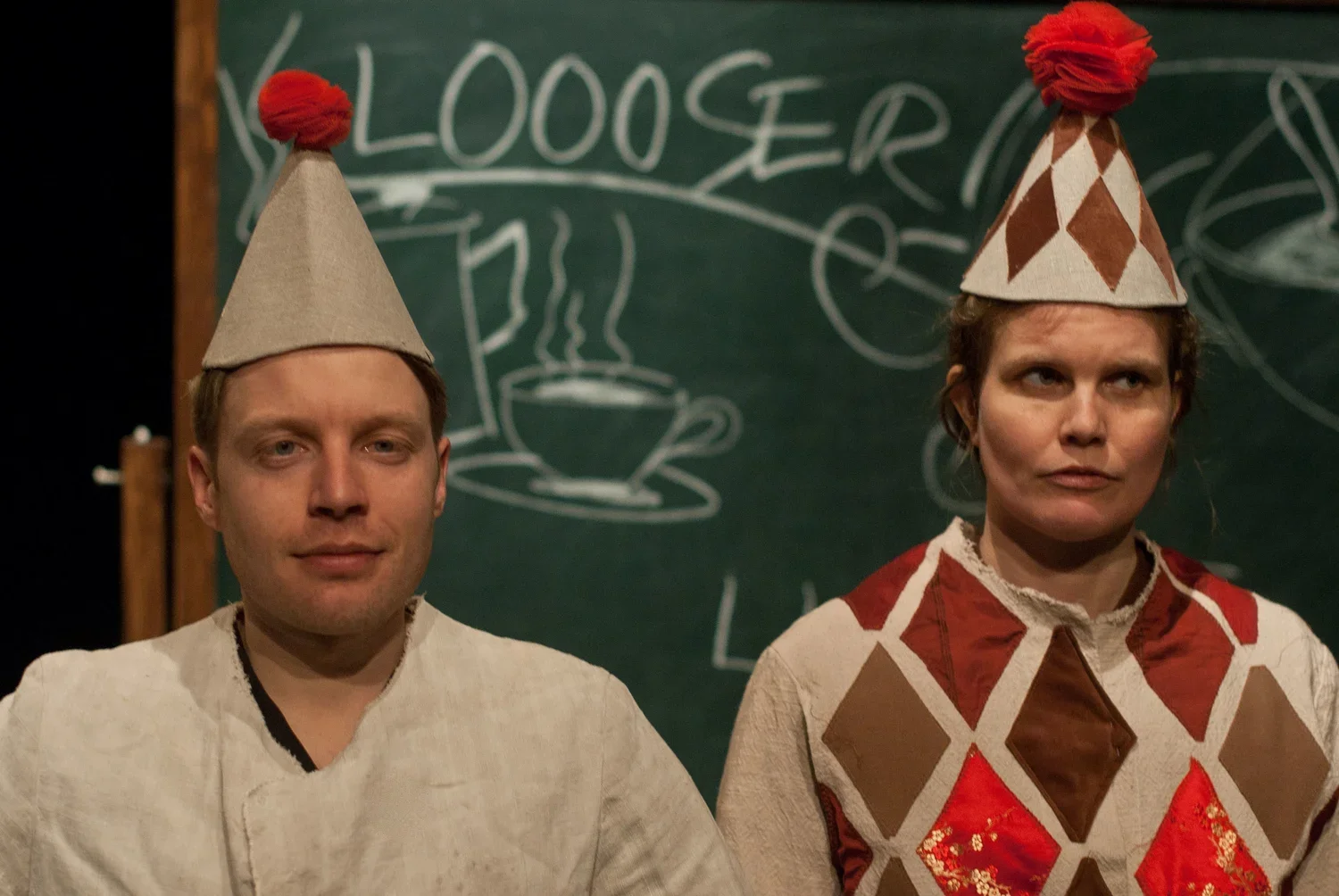 Två personer i clownkostymer och clownmössor, en man och en kvinna, framför en svarta tavla med teckningar och text.