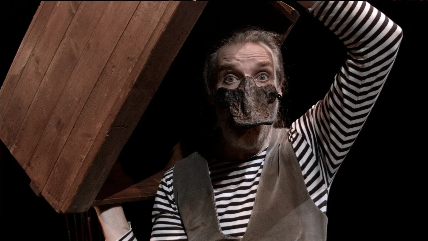 Person som bär en ansiktsmask som liknar en anknäbb, med randig tröja och ett mörkt rum, bärandes på en trälåda.