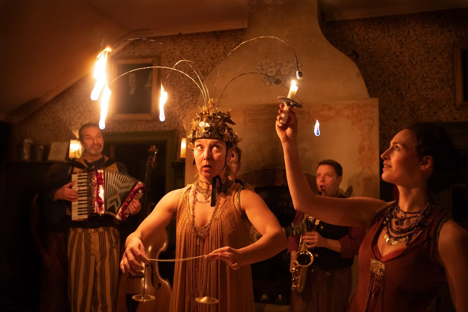 Fem personer deltar i en eldshow i ett rum, där en kvinna bär en huvudbonad med brinnande ljus, medan andra spelar musikinstrument och tittar på