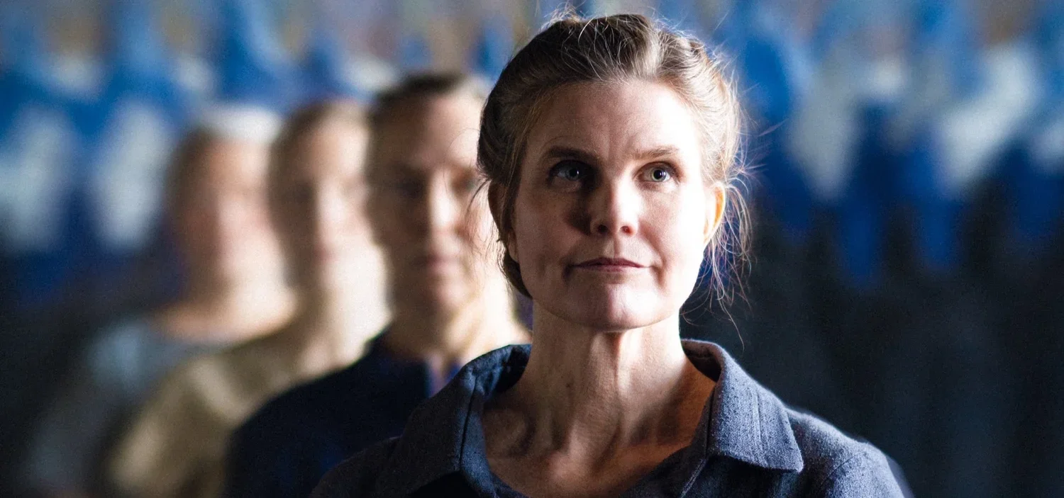 En kvinna med ljust hår i en knut, som tittar uppåt och ler svagt, på rad bakom henne står andra personer.