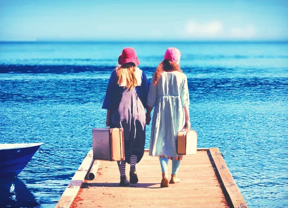 Två kvinnor i vintagekläder går på en pir mot vattnet, båda bär hatt och väskor, med havet i bakgrunden.