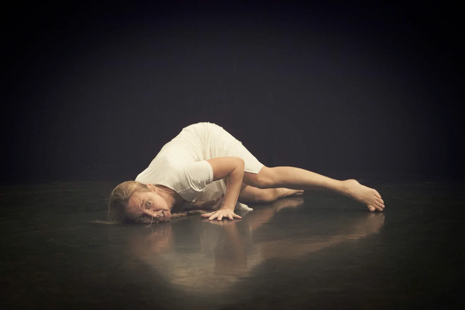 En person som ligger på golvet i ett mörkt teaterrum, klädd i vitt, med ett uttryck av rädsla eller smärta.