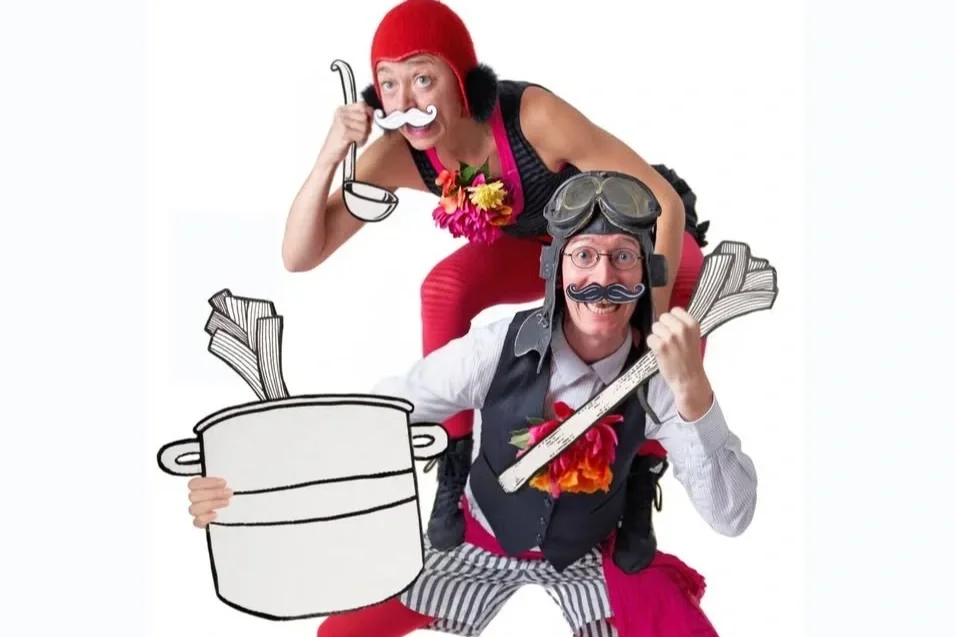 Två personer i kostymer mot en vit bakgrund, en kvinna med röd mössa håller i en tecknad soppslev, och en man med pilotglasögon, peruk och mustasch håller i ett tecknat rör av pasta.