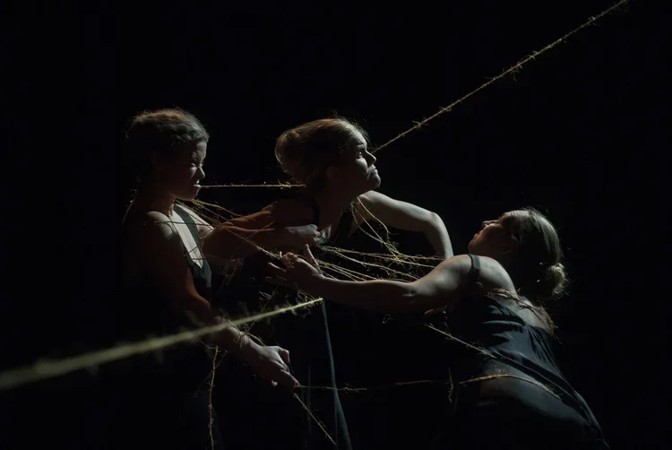 Tre personer i en teaterpjäs, arrangerade i dramatiska positioner mot en mörk bakgrund, med stråk av garn eller tråd.