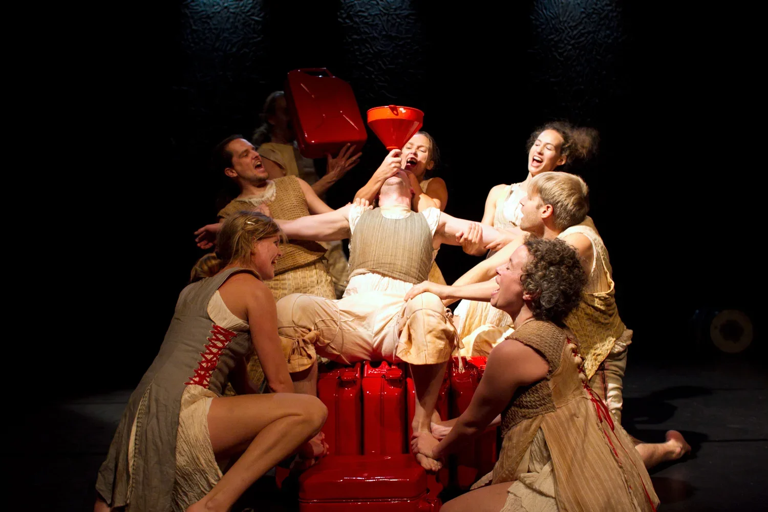 Teaterföreställning med en grupp människor i beige och bruna kostymer, som håller fast en person i mitten, som sitter på ett rött föremål med en röd tratt i munnen.