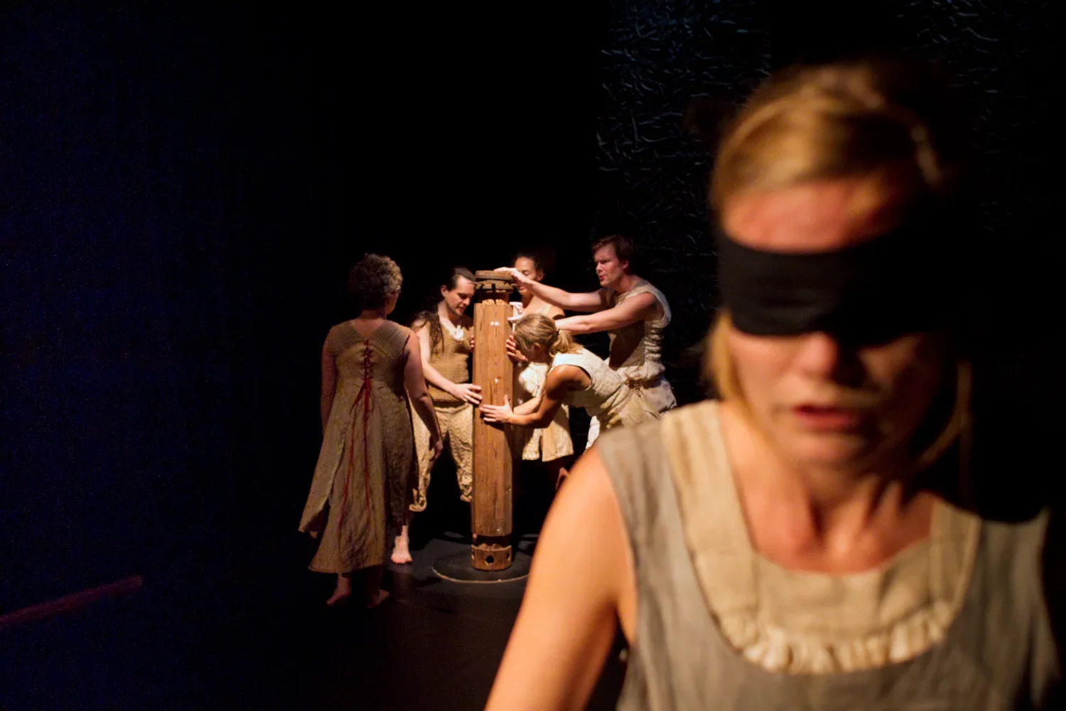 En grupp personer som deltar i en teaterföreställning, där några håller på med ett träobjekt i bakgrunden, och en person i förgrunden bär ögonbindel.