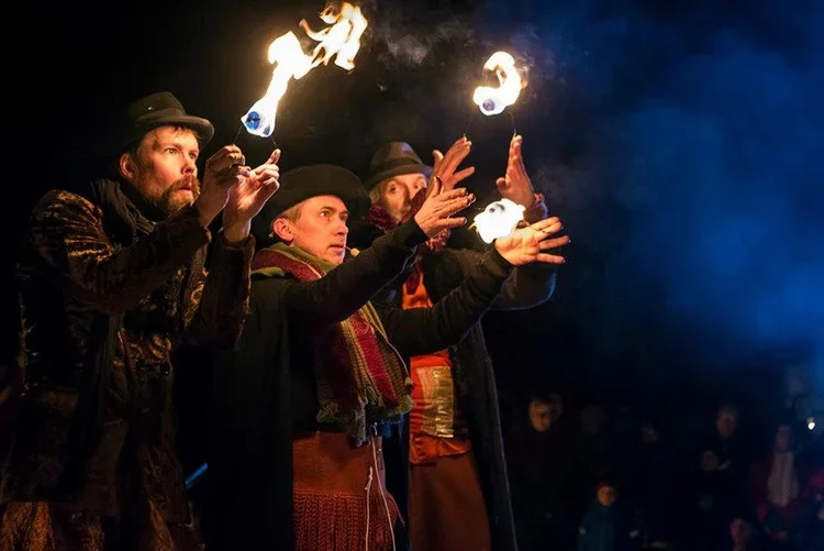 Tre personer utför en eldshow med eldklot under natten, alla iklädda kostymer och hattar, utomhus.