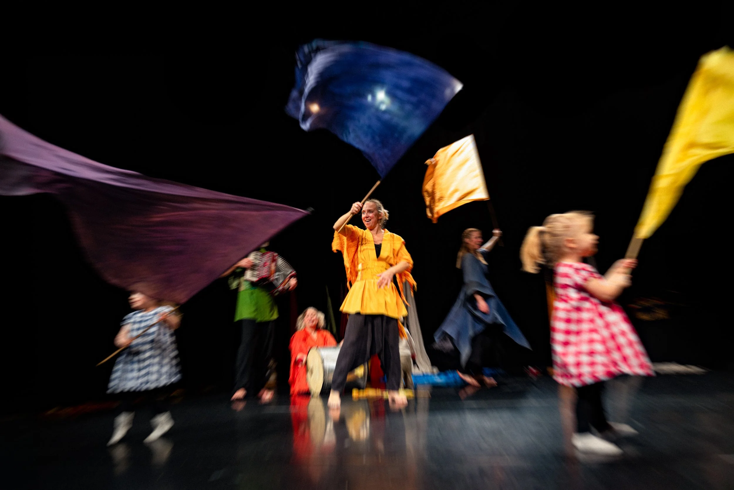 Flera människor i färgglada kläder som dansar och viftar med flaggor på en scen med svart bakgrund.