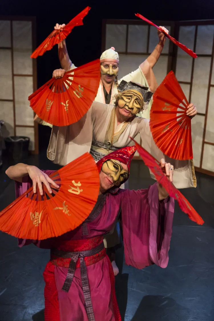 Tre personer i japanska teaterkostymer och masker som håller röda pappers- och bambu-solfjädrar, som del av en teaterföreställning.
