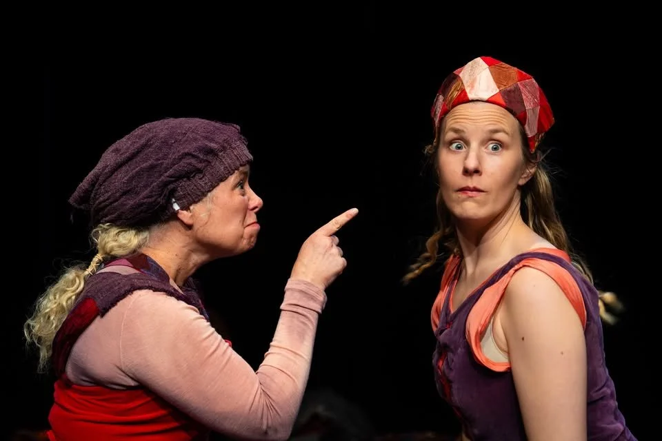 Två kvinnor i teaterkostymer, en av dem pekar finger mot den andra, som ser förvånad ut. Bakgrunden är svart.